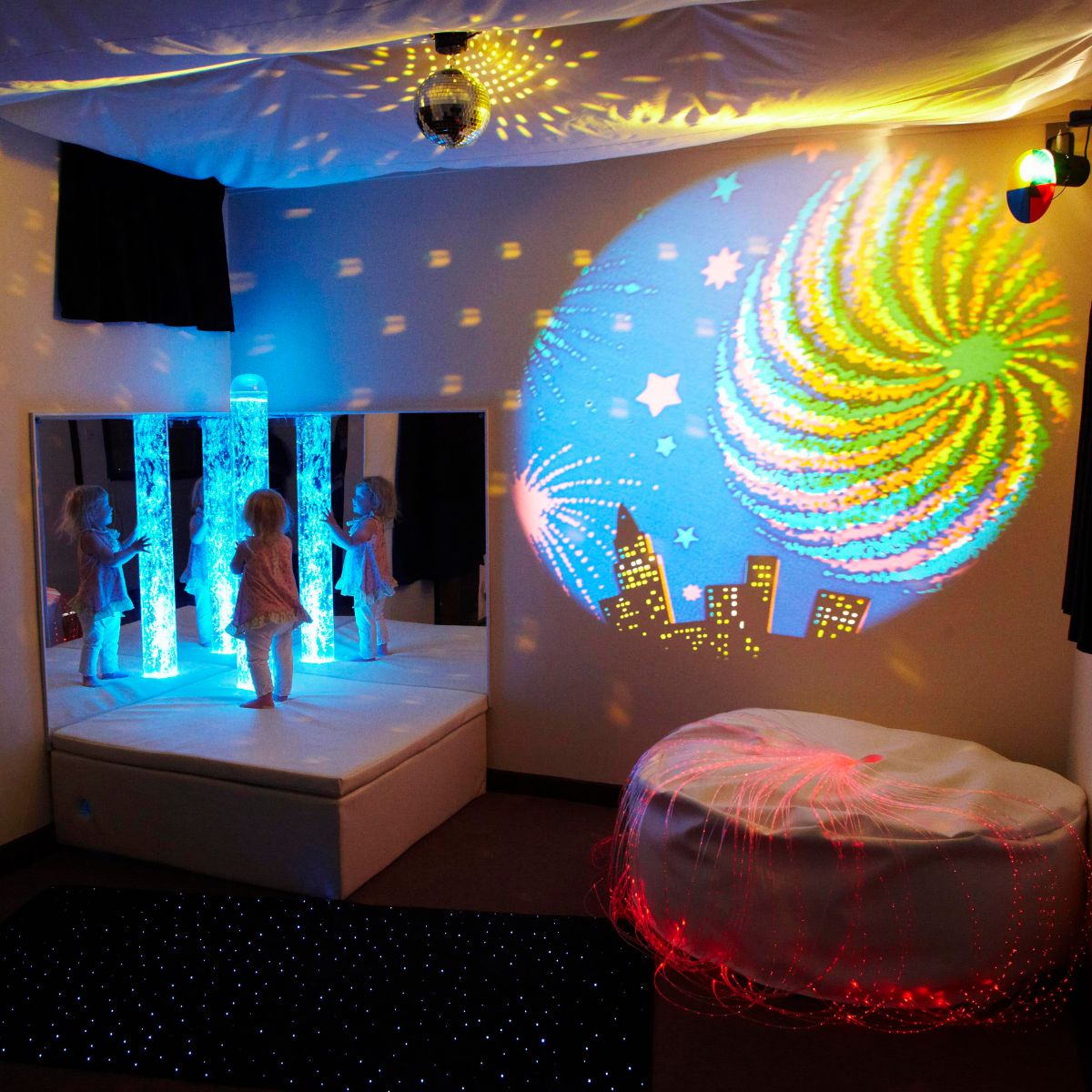 Calming Sensory Room Package with Kid standing on platform next to light blue bubble tube with red fiber optic lights and projector slide